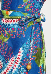 Robe portefeuille en tissu bleu avec des motifs géométriques colorés et des oiseaux, nouée à la taille par un nœud assorti avec un détail en perle en bois.