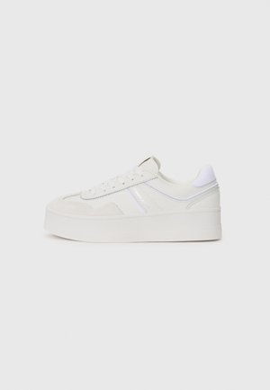 Witte lage sneaker met dikke zool, veters aan de voorkant en subtiele stikseldetails tegen een effen lichte achtergrond.