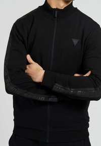 Veste noire zippée en tissu doux. Dispose d'un col montant, de poignets côtelés et du logo "GUESS" en imprimé sombre le long des manches.