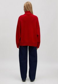 Roter gestrickter Pullover mit hohem Kragen, lässiger Passform und geripptem Saum. Kombiniert mit dunkelblauen weit geschnittenen Hosen und schwarzen Stiefeln.