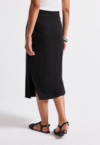 Jupe midi noire côtelée avec fente sur le côté, dotée d'une taille haute. Associée à des sandales tissées noires, mettant en avant un design simple.