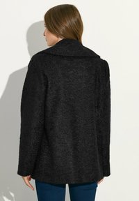 Abrigo negro de material texturizado, con un cuello amplio y mangas largas. La parte posterior es lisa con un corte relajado, que llega hasta las caderas.