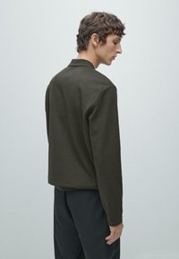 Młody mężczyzna z krótkimi, kręconymi brązowymi włosami stoi bokiem, ubrany w ciemnooliwkową kurtkę i czarne spodnie na jednolitym jasnym tle.