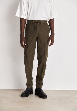 Homme portant un pantalon vert olive avec cordon, des bottes noires et une chemise blanche ample à manches courtes sur un fond uni.