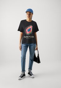 Musternaga T-särk värvilise papaya graafika ja tekstiga "PAPAYEAH ME VUELVES LOCA," koos siniste teksade ja mustade kõrgete tossudega.