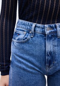 Άτομο που φοράει ψηλόμεσα μπλε τζιν παντελόνια και μαύρο μακρυμάνικο μπλουζάκι με σχέδιο, με ένα μικρό κόκκινο, λευκό και μπλε λογότυπο στην τσέπη.