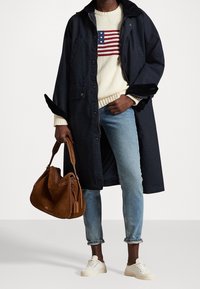 Abrigo largo azul marino, suéter crema con diseño de bandera, jeans azul claro, bolso de ante color beige y zapatillas blancas con suelas texturizadas.