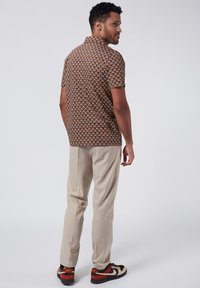 Short-sleeve shirt met een bruin geometrisch patroon, gecombineerd met beige broek en zwart/rode sneakers, gezien vanuit de achterkant.