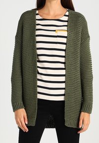 Cárdigan de punto verde oliva con frente abierto, tejido texturizado y mangas largas, usado sobre una camisa a rayas con un bolsillo amarillo.