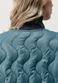 Giacca imbottita teal trapuntata con motivo a onde, dotata di un colletto alto nero e texture liscia. La cucitura visibile aggiunge dettagli.