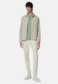Giacca grigia chiara con due tasche sul petto sopra una camicia verde chiaro, abbinata a pantaloni bianchi e sneakers bianche. Tessuto liscio, design casual.