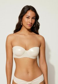 Sujetador sin tirantes beige con una textura lisa y brillante y soporte con aros. Presenta un diseño sencillo con detalles de costura horizontal.