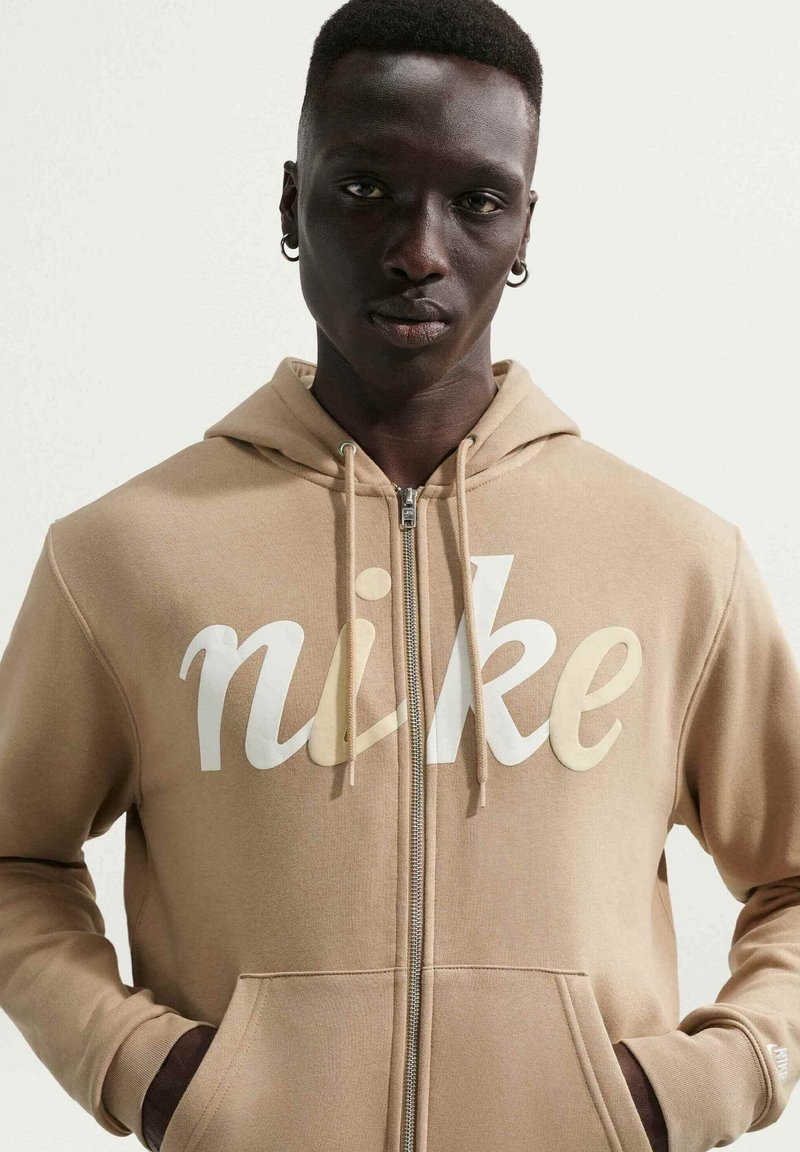 Man wearing a tan zip-up hoodie with "nike" in large white and beige letters, hands in pockets, standing against a plain light background.