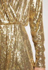 Robe longue à manches longues en sequins dorés avec corsage style cache-cœur et taille cintrée, montrant la partie médiane et le bras droit.