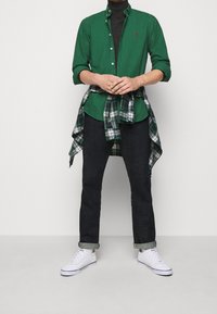 Grön knapp-up skjorta med uppvikta ärmar, svart polotröja, mörka jeans och vita sneakers. Rutigt flanell knutet runt midjan.