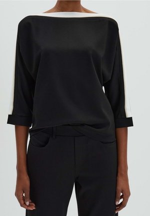 Kobieta w czarnym topie z dekoltem w łódkę z białymi paskami na rękawach oraz czarnych spodniach, stojąca na jednolitym tle.