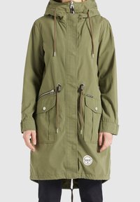 Parka à capuche vert olive avec fermeture éclair et poches à bouton-pression, cordons de serrage réglables et patch circulaire de marque sur l'ourlet inférieur avant.