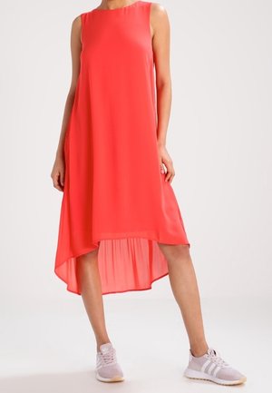 Kobieta w koralowej, bezrękawnikowej sukience z asymetrycznym dołem, w jasnoróżowych trampkach, stojąca na tle jednolitego tła.