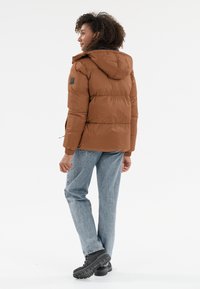 Brun isoleret pufferjakke med hætte, der har en tekstureret overflade, elastiske manchetter og et logo-patch på venstre arm, båret med blå jeans.