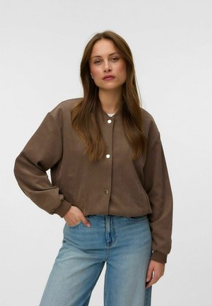 Vrouw met lang bruin haar, die een bruin gebuttoned jasje en lichtblauwe spijkerbroek draagt, staand met één hand in de zak tegen een effen achtergrond.