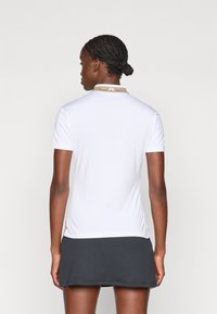 Bela polo majica s smetano ovratnikom, kratkimi rokavi in prilegajočim krojem, v kombinaciji s temno naborkano krilo. Gladka tekstura, minimalno označevanje.