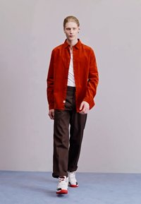 Tenue masculine composée d'une chemise boutonnée en velours côtelé orange, d'un t-shirt blanc, de pantalons marron et de baskets avec des accents rouges sur un fond clair.
