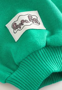 Tessuto di maglione verde a maglia con polsini a coste. Presenta un'etichetta bianca con "Peppa Pig" in caratteri rosa. Texture liscia, design informale.