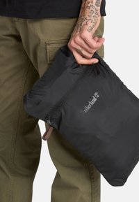 Svart nylonfodral med dragkedja, prytt med en Timberland-logotyp. Det har en strukturerad yta och en brun tygdetalj vid dragkedjehandtaget.