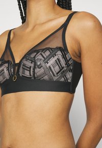 Black bralette with mesh and geometric embroidery, featuring an underband, adjustable straps, and a gold accent at the center front.