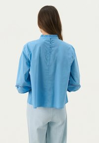 Blusa azzurro chiaro, morbida e ampia, con colletto arricciato corto, chiusura con bottoni sulla schiena e bordi in pizzo sulle maniche. Tessuto di cotone morbido.