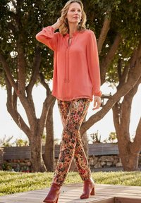Pfirsichfarbene Bluse mit Schlüsselloch-Ausschnitt und Kordelzug, kombiniert mit floralen Hosen in erdigen Tönen und braunen, knöchelhohen Stiefeln mit Absatz.