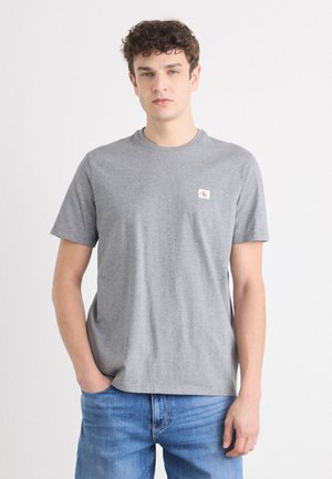 Ung mann med krøllete hår iført en grå kortermet T-skjorte med liten brystlogo og blå jeans, stående mot en hvit bakgrunn.