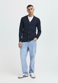 Marinblå cardigan med V-ringning och sex knappar, strukturerad tyg, i kombination med ljusblå jeans och vita sneakers med svarta ränder.