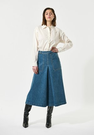 Denim-Maxi-Rock mit Vorderteil-Details, das eine ausgeschnittene Silhouette und eine hohe Taille aufweist. Kombiniert mit einem hellen, knopfbaren Hemd.