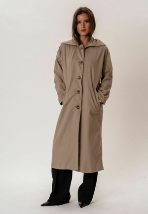 Kobieta w długim beżowym płaszczu na guziki z szerokim kołnierzem, ręce w kieszeniach, w połączeniu ze szerokimi czarnymi spodniami i czarnymi, spiczastymi butami.