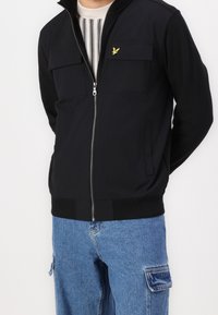 Chaqueta negra con cremallera y dos bolsillos en el pecho, combinada con pantalones cargo de denim azul claro. Presenta un pequeño logo amarillo en el pecho.