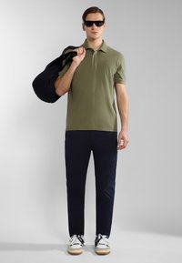 Polo shirt verde oliva con colletto, pantaloni blu navy e zaino nero. Occhiali da sole neri e sneaker bianche con accenti navy.