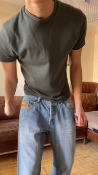 Άτομο που φοράει σκούρο πράσινο μπλουζάκι και ανοιχτά μπλε τζιν, στέκεται μέσα σε εσωτερικό χώρο κοντά σε καφέ καναπέδες σε ξύλινο πάτωμα.