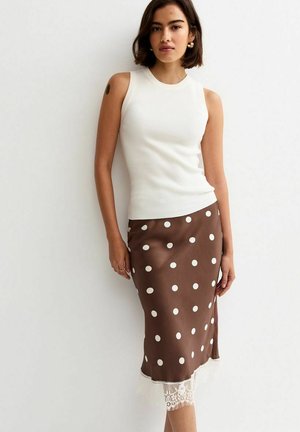Kobieta ubrana w białą, bezrękawną prążkowaną bluzkę i brązową spódnicę midi w duże białe kropki z koronkowym wykończeniem, stojąca na tle białej ściany.