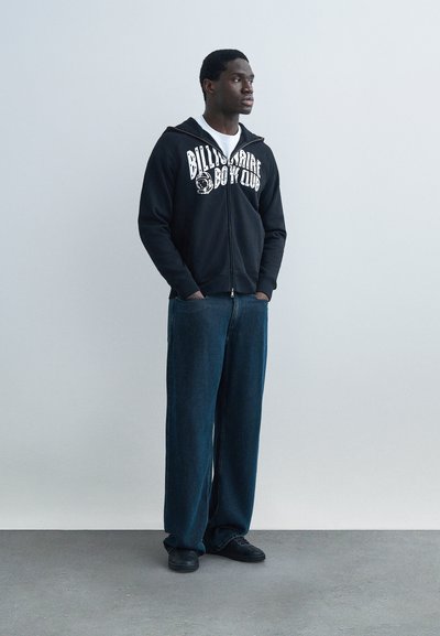 Νεαρός άνδρας στέκεται με τα χέρια στις τσέπες, φορώντας μαύρο φούτερ με φερμουάρ και λευκό κείμενο «Billionaire Boys Club» και χαλαρά μπλε τζιν.