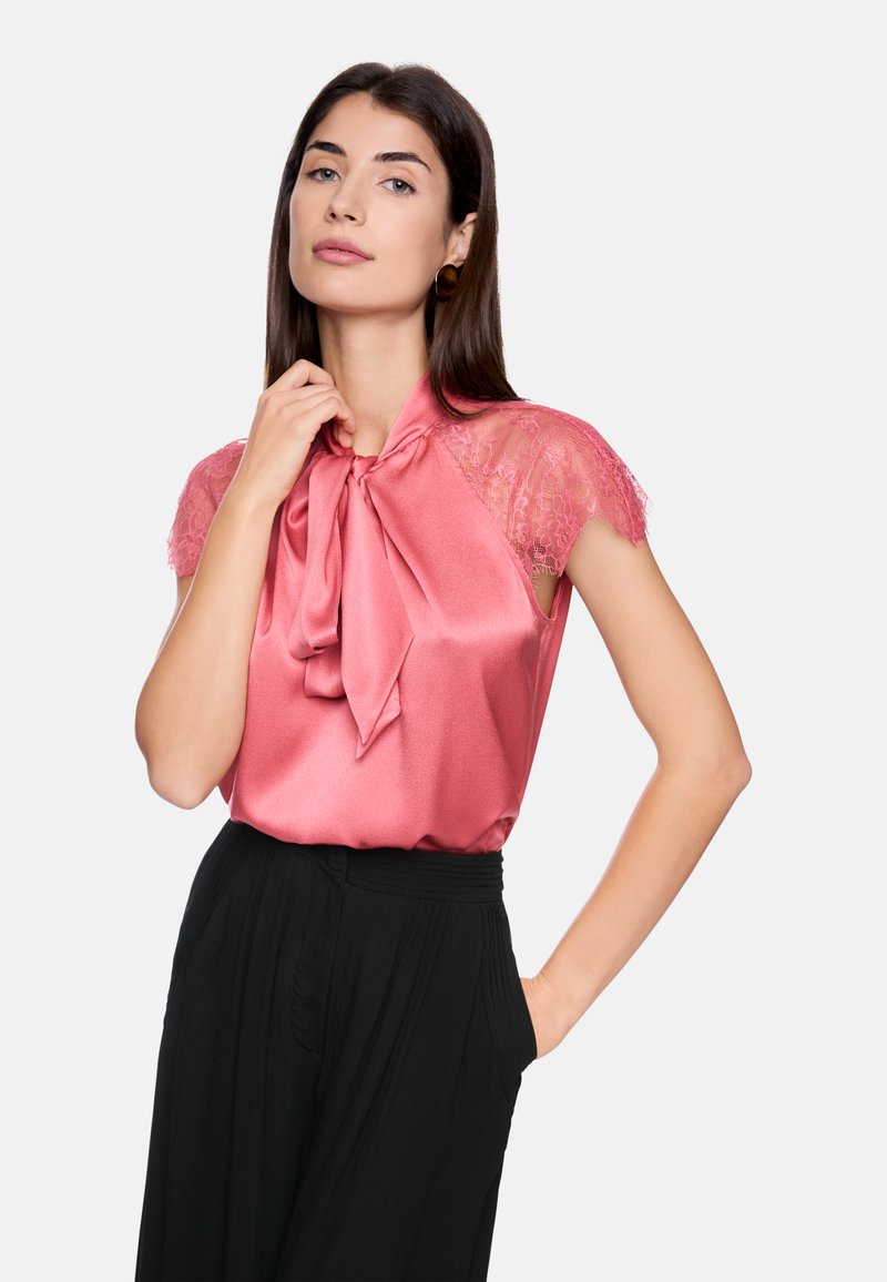 Rosa Satinoberteil mit einer Schleife am Hals und Spitzenärmeln, kombiniert mit schwarzen, hochgeschnittenen Hosen. Glatte Textur, elegantes Design.