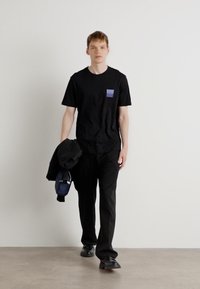 T-shirt en coton noir avec une petite patch violet, associé à un pantalon noir et des chaussures noires. Une veste noire est tenue dans la main gauche.