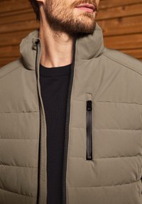 Chaqueta acolchada en verde oliva con un cuello alto, cremallera frontal y un bolsillo en el pecho con cremallera. Lleva puesta sobre un suéter negro de tejido texturizado.