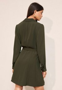 Rochie petrecută verde închis cu mâneci lungi, croială ajustată și talie evidențiată. Are guler și fustă plisată. Textură netedă a materialului.