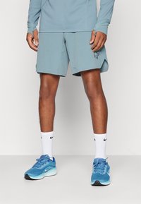 Ανοικτό μπλέ αθλητικά σορτς με χαλαρή εφαρμογή, διαθέτοντας κεντητό λογότυπο. Συνδυασμένα με λευκές κάλτσες και μπλε αθλητικά παπούτσια.