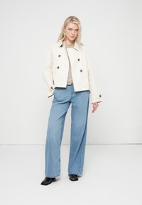 Veste courte couleur crème avec de gros boutons marron, associée à un jean bleu clair à jambes larges. Le modèle est debout devant un fond neutre.