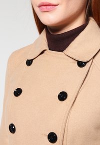 Abrigo de lana beige con un diseño de doble botonadura, con botones redondos negros, un cuello doblado y un cuello alto de canalé marrón debajo.