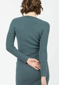 Ribbad långärmad klänning i mörk teal. Tajt silhuett med rund ringning. Texturen verkar mjuk och elastisk, vilket framhäver kroppens form.