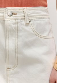 Jean en denim blanc avec des coutures contrastées et une fermeture à bouton argenté, dotés d'une poche avant et d'une texture lisse.