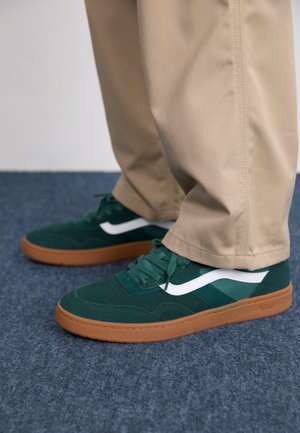 Adidași din suede verde cu accente albe și o talpă de cauciuc maro, având șireturi plate și un design low-profile.